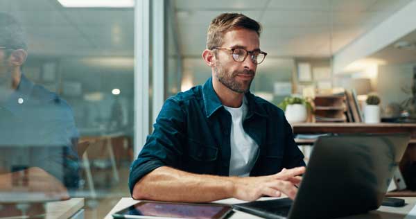 man wearing glasses working on laptop in office situation, reflection on glass divider to left